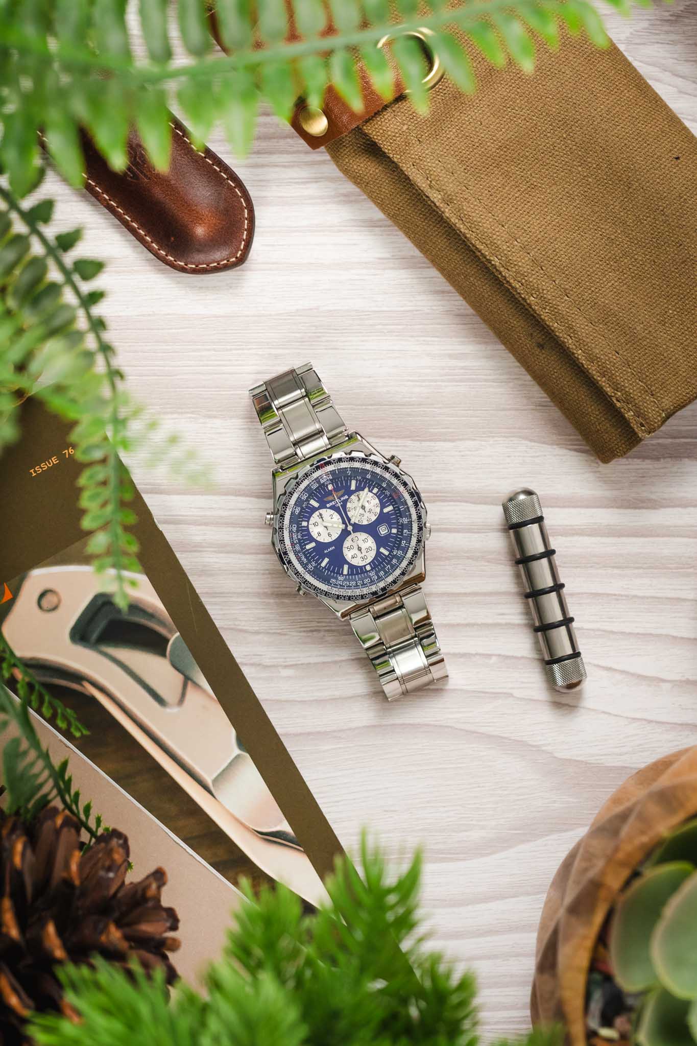 An overhead lifestyle photograph of a Breitling Jupiter Pilot watch centered on a light wood grain surface, framed by green fern leaves and travel accessories.