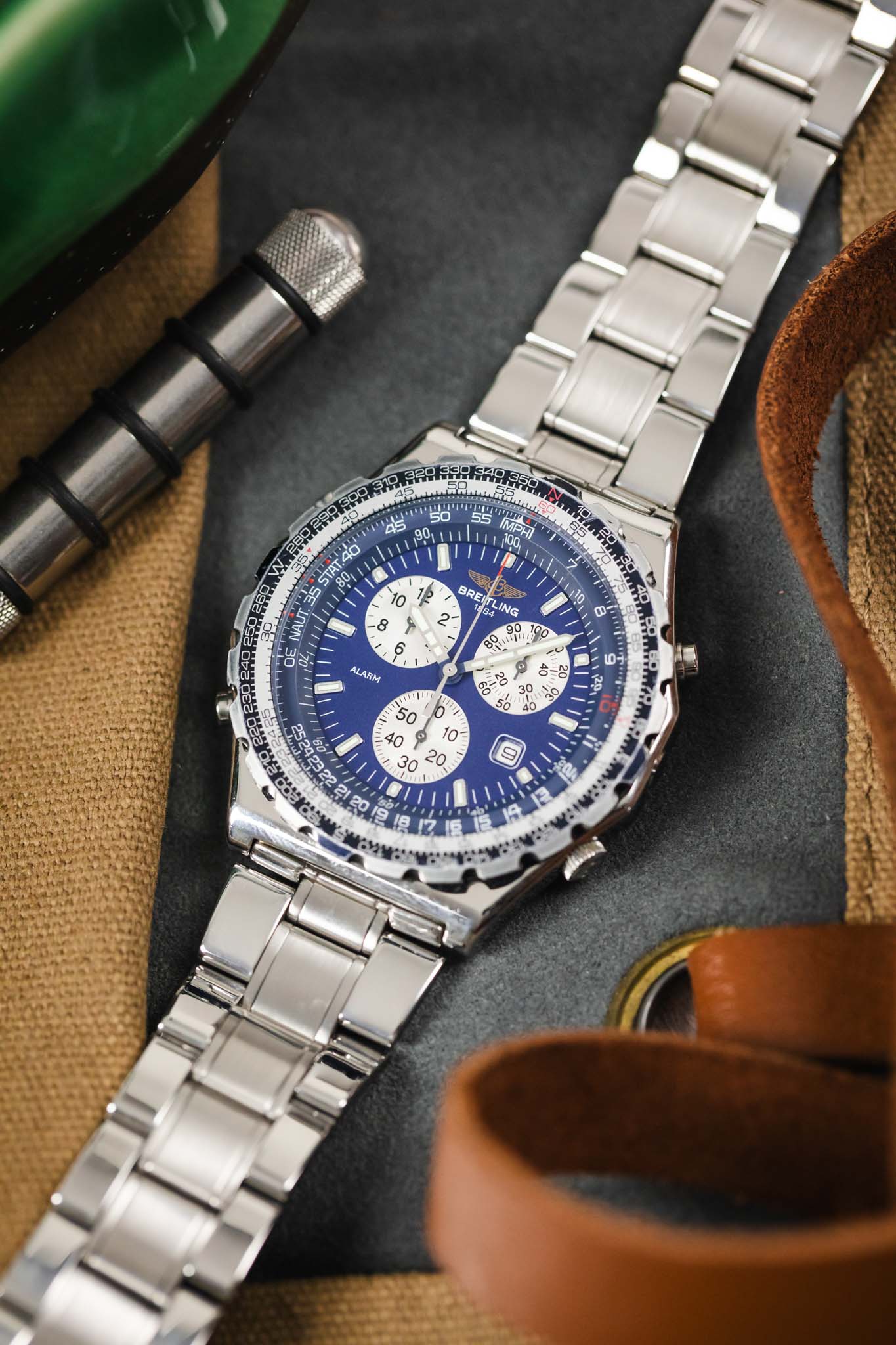 A top-down lifestyle shot of a Breitling Jupiter Pilot watch laying on a grey and tan surface next to a metal spring bar tool and leather straps.