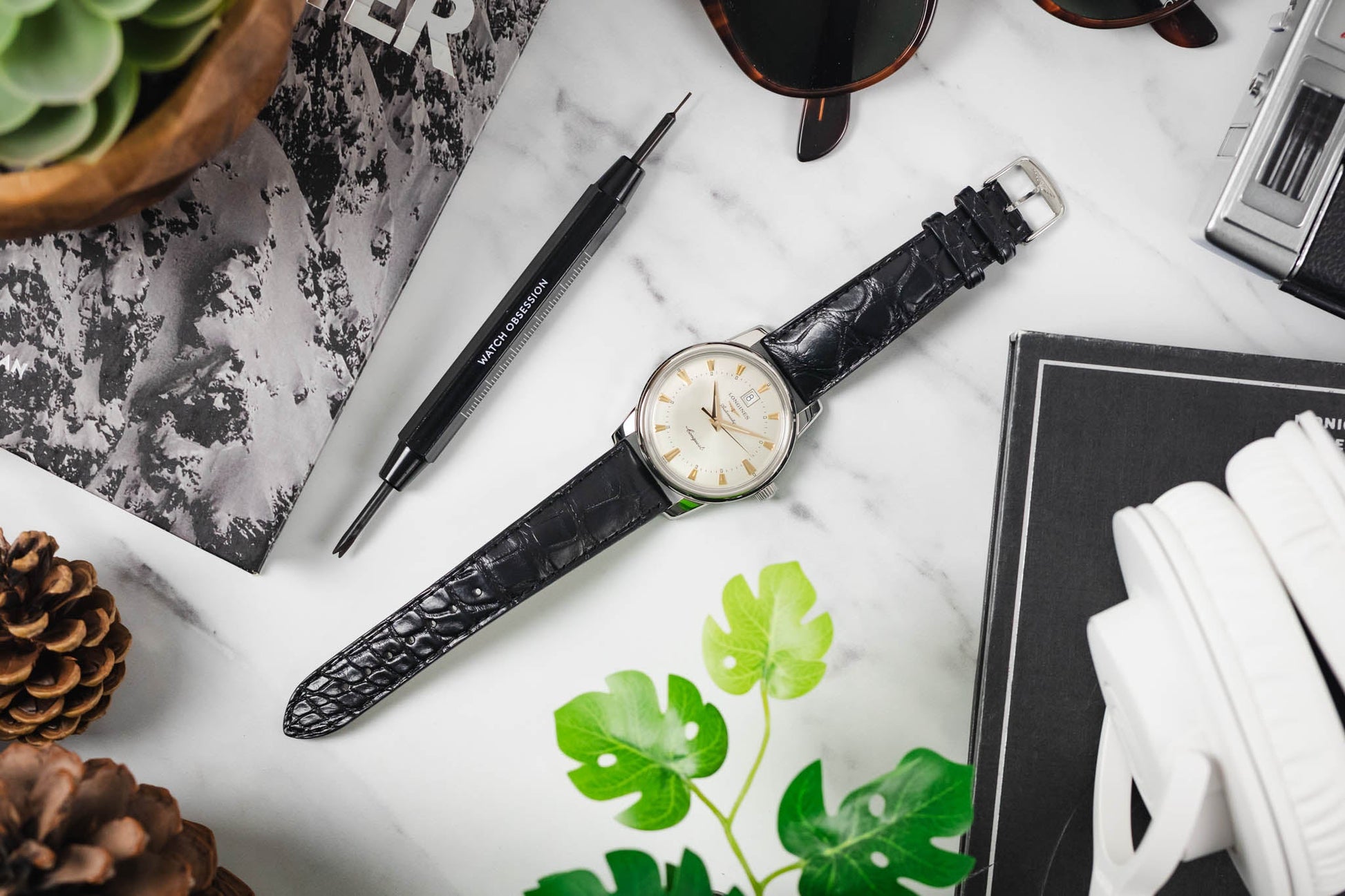 Black longines watch wristwatch with white face on a marble surface with plants and stationery items.