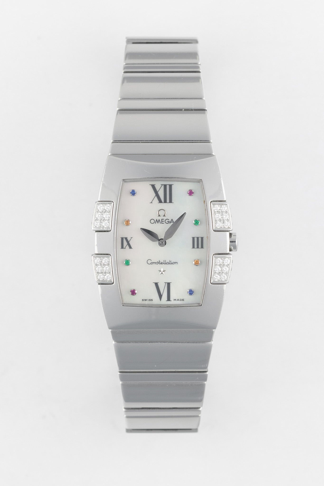A top-down studio shot of a rectangular Omega Constellation Quadra watch with a white mother-of-pearl dial and a stainless steel integrated bracelet. The dial features multi-colored gemstone hour markers and diamond-set "claws" on the case sides.