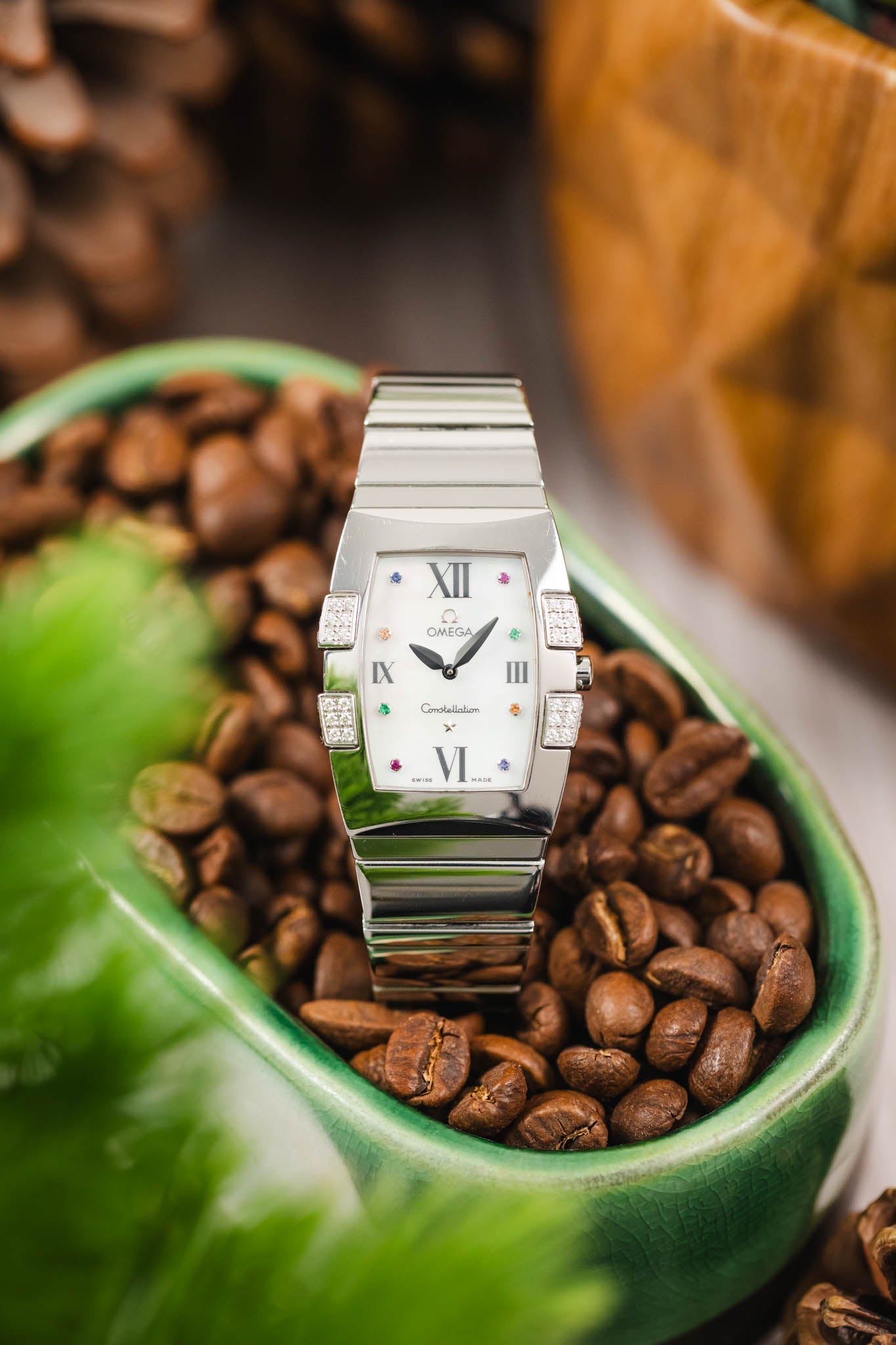 A creative lifestyle shot of the Omega Constellation Quadra standing upright inside a green bowl filled with roasted coffee beans, with blurred green foliage in the foreground.