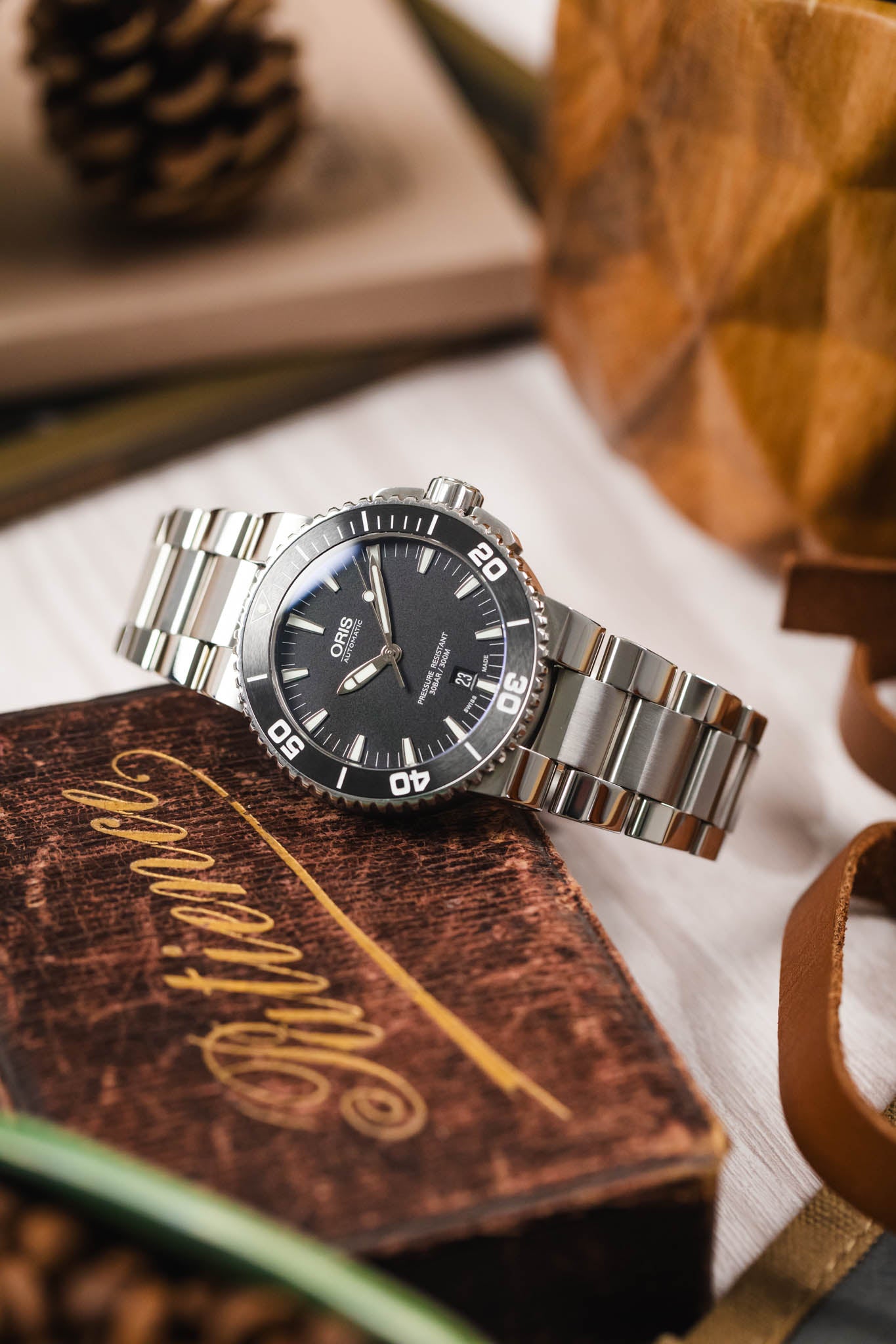 A lifestyle image of an Oris Aquis Date watch with a steel bracelet resting on a vintage brown book titled "Patience," surrounded by leather accents and out-of-focus greenery.
