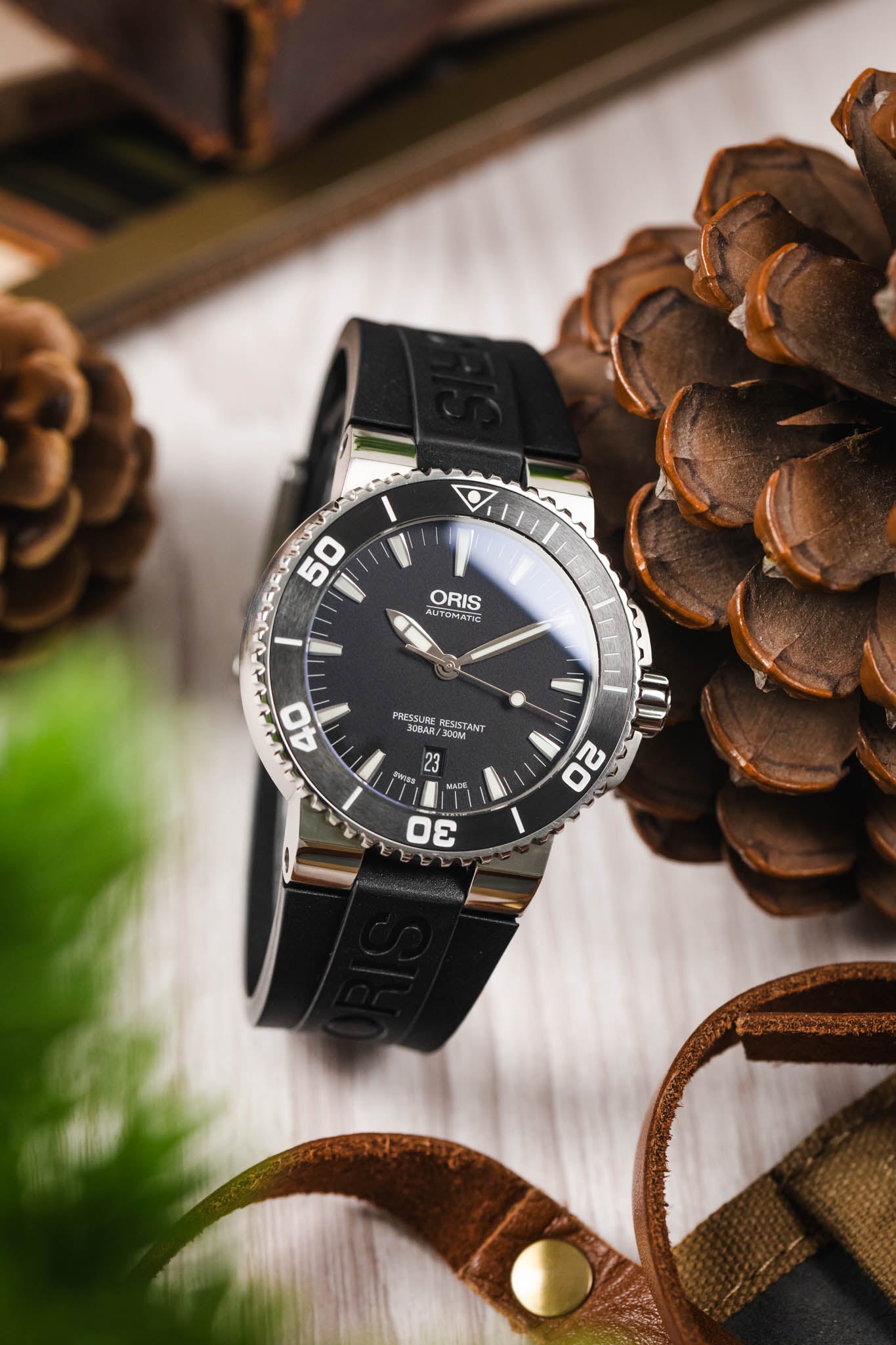 A lifestyle photograph of an Oris Aquis Date watch with a black dial and black rubber strap, positioned next to a large brown pinecone and a leather strap on a light wood surface.
