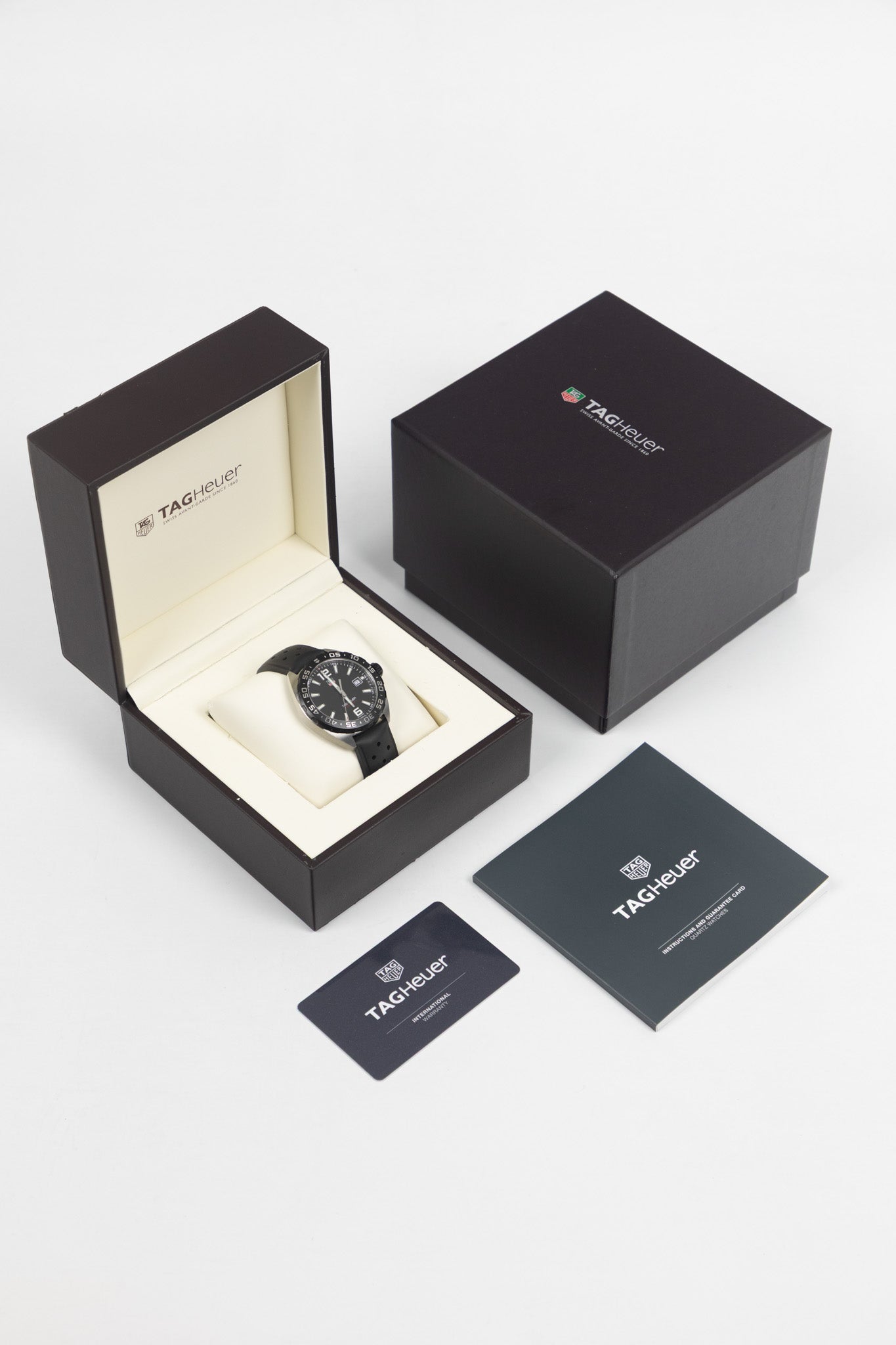 A full set display including the watch on a white cushion inside its open brown presentation box, the outer box lid, an instruction manual, and an international warranty card.