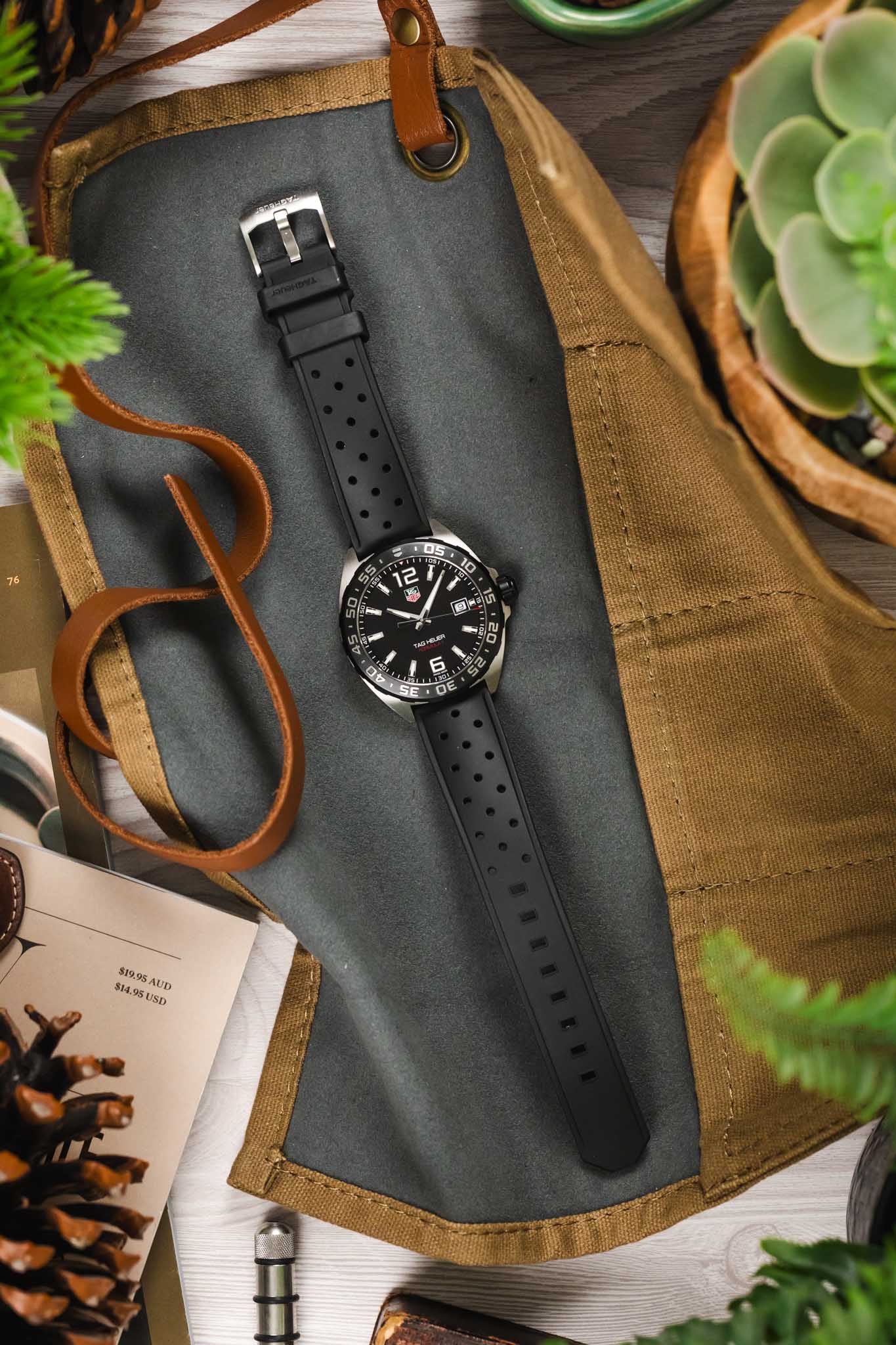 A top-down lifestyle shot of a TAG Heuer Formula 1 watch with a black dial and black perforated rubber strap, resting on a grey fabric inside a tan canvas tool roll.