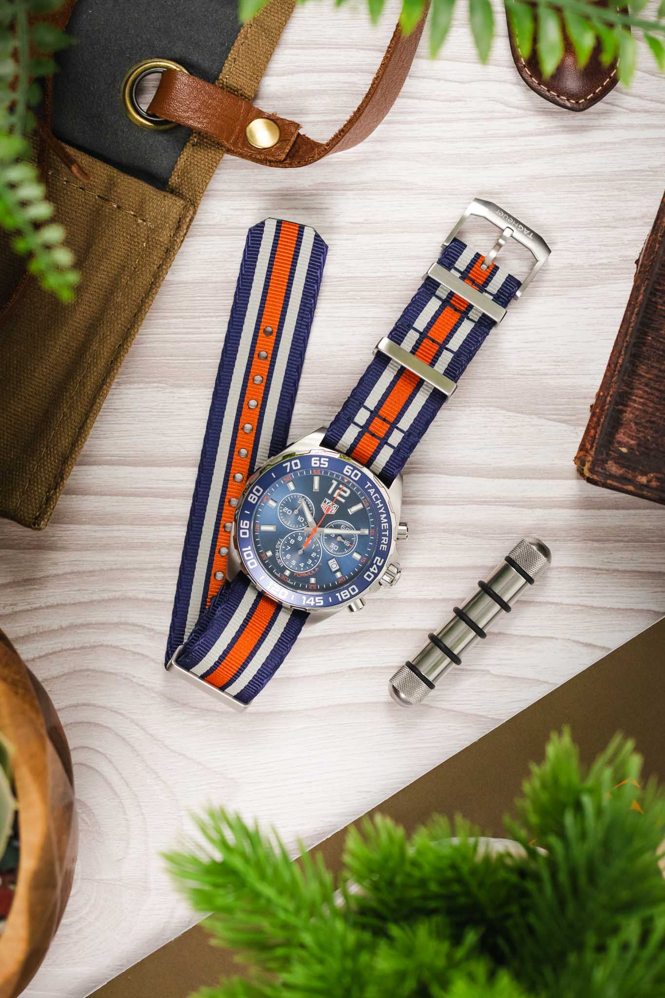A flat-lay shot of the watch with its blue, orange, and white striped fabric strap resting on a light wood surface next to a tan canvas roll and a steel spring bar tool.