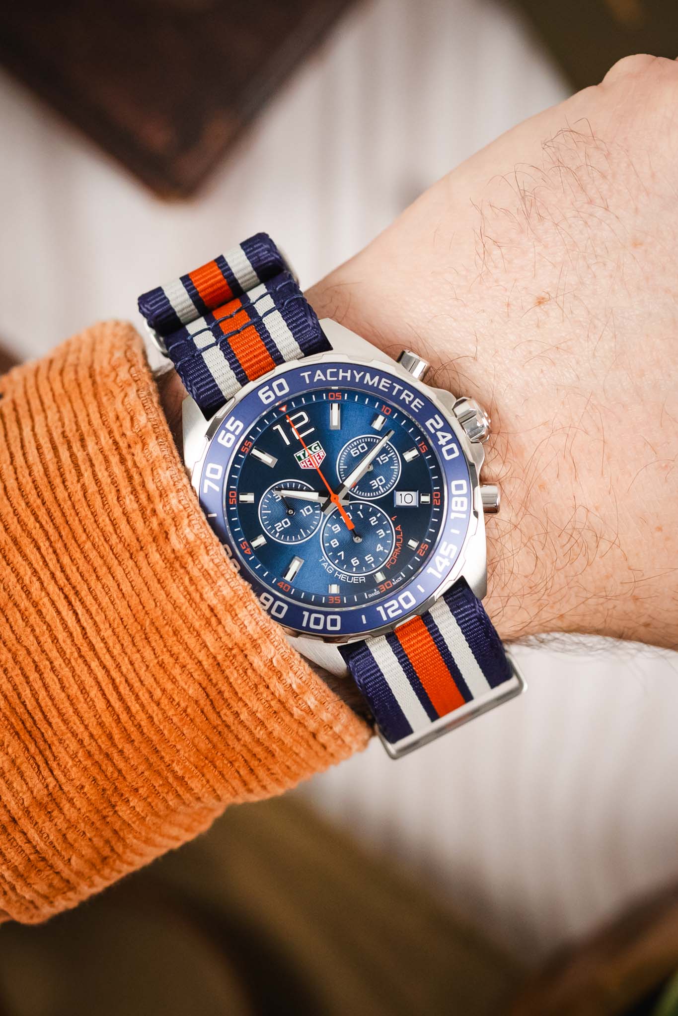 A close-up lifestyle image of the watch on a wrist, worn with an orange corduroy shirt, highlighting the vibrant orange and blue dial details.