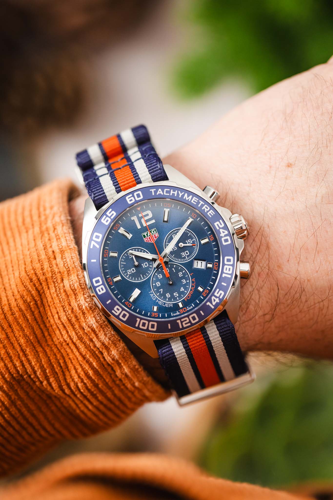 An angled wrist shot showing how the striped fabric strap and blue tachymeter bezel complement a casual autumn-colored outfit.