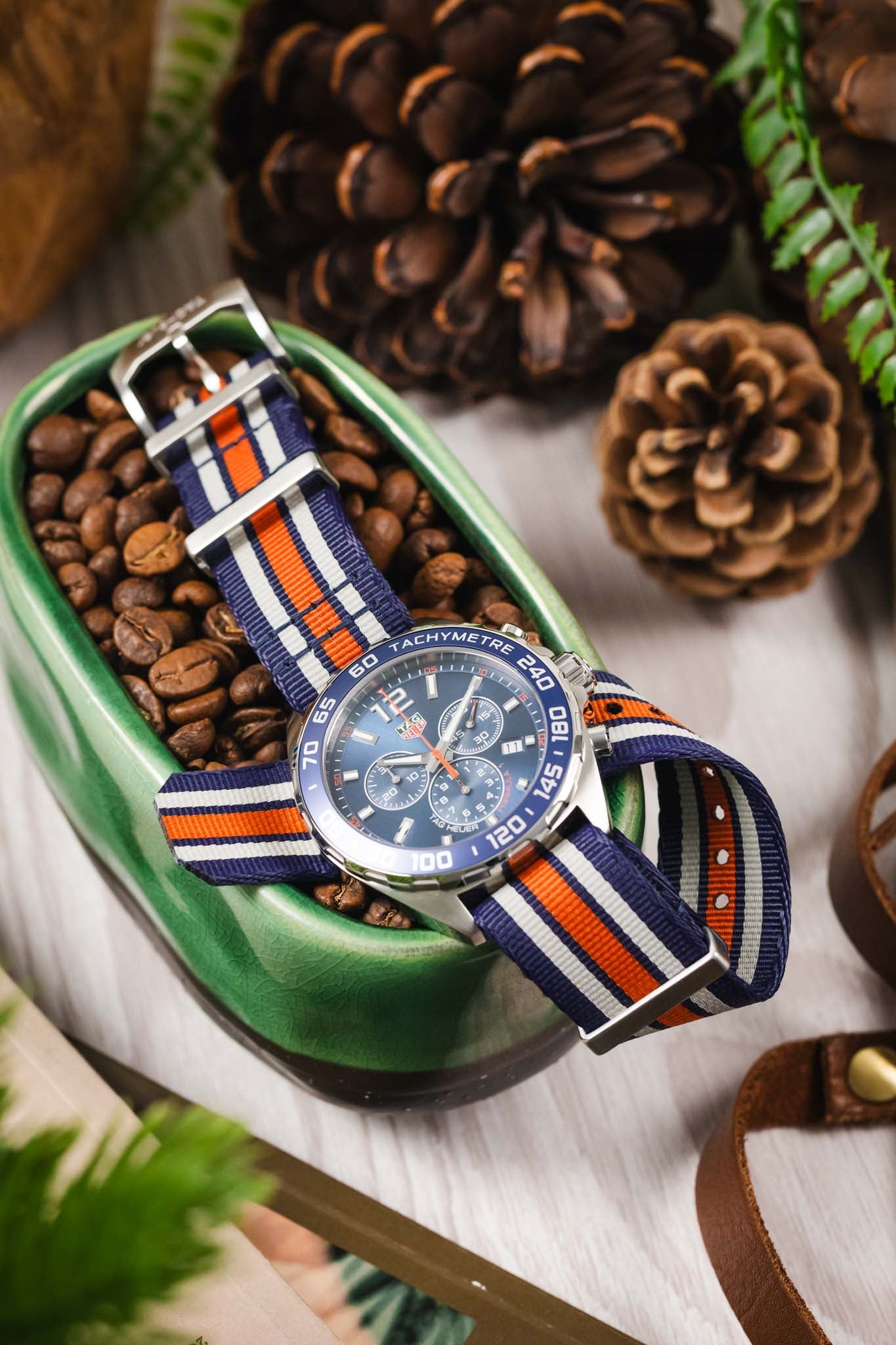A creative overhead shot of the Gulf Edition watch resting in a green ceramic bowl filled with dark roasted coffee beans, surrounded by pinecones.